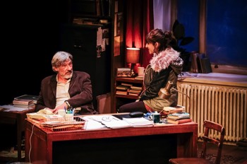 Frank (Simon Armstrong) sits at his desk with Rita (Taj Atwal) perched on the corner of the desk. It's dark outside the large sash window. Frank's academic office is lit with various lamps. There are papers neatly organised across his desk, his glasses lay on top of an open document.