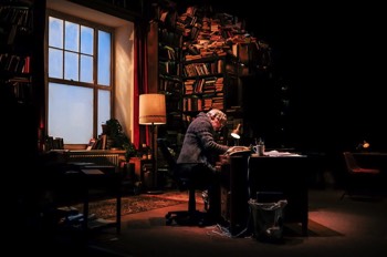 Frank (Simon Armstrong) sits at his desk writing. He's surrounded by bookshelves. Afternoon sun streams in through the large window, but the room is fairly shadowy. The small desk lamp beside him is switched on.