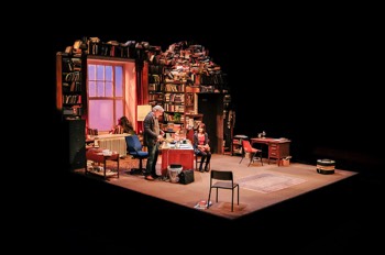 A wide angled photo of the entire stage. Frank (Simon Armstrong) stands behind his desk in his academic office. Early evening sunlight streams through a large sash window in a wall which is mostly just books. Above the doorway is a cascading pile of books which continues down the walls.
