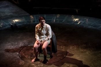 Kay Langrish (Jodie McNee) sits in a chair in the centre of a circular stage. The central area is charcoal grey with some floral floor patterns visible in places, then surrounded by a ring of corrugated metal. She wears a dull white shirt and her hiar is slicked back. A jacket hangs over the back of her chair along with her trousers. She holds a cigarette in her hand.