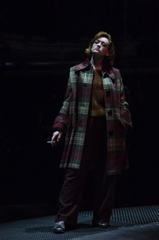 Julia Standing (Lucy Briggs-Owen) stands in the dark wearing a long tartan jacket. She looks up into light from above. A cigarette smokes in her hand to her side.