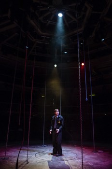  Kay Langrish (Jodie McNee) stands in the centre of the circular stage in a single soft cold spotlight. There is the red glow from lights suggesting she is surrounded by fire. Thick chains hang down from the top of the theatre, along with military grade cabling. 