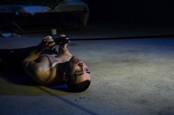  Bobby (Jake Davies) lays on his back on the stained beige carpet holding a Playstation controller to his chest, the cable of which is pulled tight and extends out of the photo to the right. He cranes his head baack into the light of the television out of frame. Otherwise, the whole stage is in darkness. 