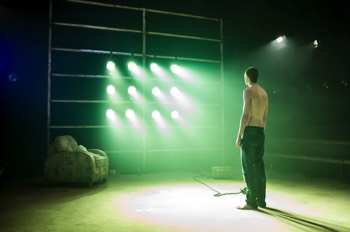  Hench (Alex Austin) stands staring into a wall of lights symbolising a television screen fixed to a large climbing frame style wall. The lights emit a green-ish colour bright light. The rest of the stage is dark. 