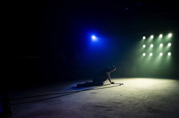  Hench (Alex Austin) lays on the floor in the centre of the stage. A tall bank of lights shine dimly at the end of the space. A single blue light shines behind him. There's a long black rope to his side. 