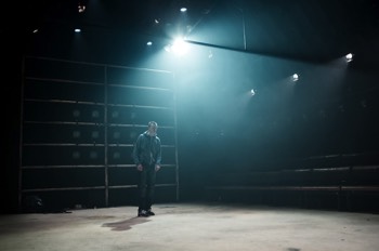 A wide angles shot along the traverse stage with Hench (Alex Austin) stood in the centre. He's wearing a thick grey hoodie over jeans. A bright cold light fills the stage with light, suggesting a cold outside day.