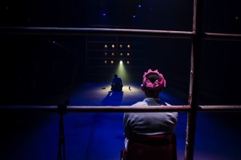 A wide angles shot along the traverse stage. Bobby (Jake Davies) is sat closest to the camera wearing a paper Christmas cracker hat with his back to the camera. Beyond him is the wall of lights symbolising the television. Hench (Alex Austin) is sat on the floor in front of the lights with one light slightly brighter creating a pool of light around him. He sits fancing the lights.
