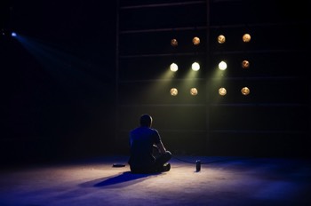  Hench (Alex Austin) sits on the floor in front of a wall of lights symbolising a television. The room is dark but three lights on the wall are slightly brighter than the others lighting him sat on the floor playing playstation. 