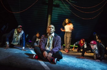 Andy (Bradley Cumberpatch) sits cross legged at the front of the image in a cold moonlight. The rest of the cast sit in the darkness staring at him. Eva (Cristal Cole) stands behind in a bright warm light looking annoyed with him.