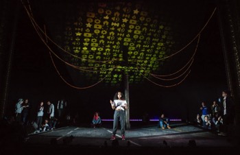 Eva (Cristal Cole) stands in a bright white light in the centre of the stage. Above her, yellow coloured emojis cover the large cantilevered ceiling.