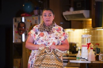  Paige (Ashley McGuire) is stood at the end of the kitchen counter holding a large zigzag fabric handbag. She's wearing a floral dress and a checked silk scarf. The blender filled with strawberry shake is to her left, as are two squirty water bottles should she need them. The large fridge in the background is out of focus, but covered in many magnetic letters and a cut-out mask of Obama. 