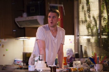  Isaac (Arthur Darvill) stands at a kitchen counter pressing his arms down on to the worktop by his sides. The countertop is strewn with prescription bottles, a blender, shake powder, a jar filled with colourful straws and a trans-flag mug. There's a window to his left with sunlight streaming through, the shadows of a plant are on the window. 