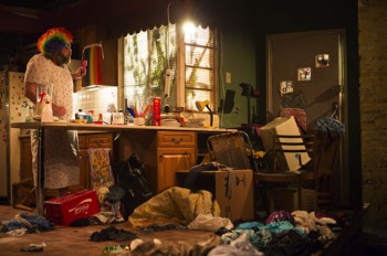 The image looks like a naturalistic home interior. There's sunlight shining through a window with some foliage behind, and metal venetian blinds. There are mounds of hoarded objects everywhere, and stuff strewn across the floor. The front door to the right is completely blocked by chairs, boxes, an ironing board and an overhead projector on a trolley. Someone is visible through the small windows at the top of the door attempting to open the door. There's a kitchen counter extending out into the space, covered in more mess. Arthur (Andy Williams) a fifty year old man is stood in a floral nightgown behind the counter licking a spatula. He's wearing a rainbow coloured clown wig and has some messily applied make up on his face. There's some gay pride stickers on the front door and a pride flag sellotaped to a cupboard door.