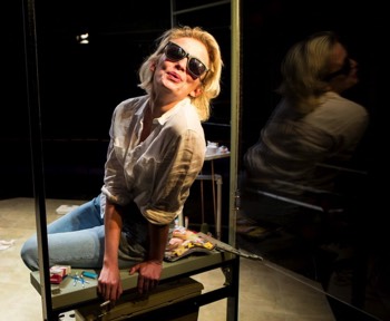 Mary (Sophie Melville) leans out an open window into the sunlight blowing out cigarette smoke. She wears black plastic sunglasses. The window is directly above the avocado toilet, which is visible thru