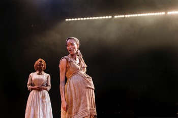 Grace (Cassie Clare) stands in very heavily worn overalls, to look like a plantation slave. All the material of her outfit is fraying and stained. She is pregnant. Dido (Emmanuella Cole) is dressed in finer clothing as a slave who works in the plantation house. A long line of lights glow behind them.