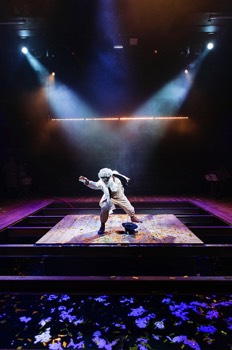 Pete (Alistair Toovey) dressed as a male slave, wearing black-face paint which was applied during the start of the show, stands in the middle of a small platform over a now water filled pit. White and gold confetti are floating on the water's surface. Pete is in two bright white spotlights, whilst the rest of the space is lit in a dark blue.