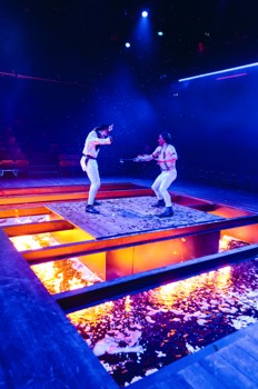 M'Closky (Ken Nwosu) dressed as a white land owner in white-face is fighting Wahnotee (Kevin Trainor) who is dressed as a native American wearing red-face. The fight on a small platform over a pit filled with flaming water. They are lit in dark blue and the warmth of the flames lights them and the pit.