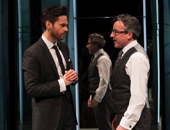  Seth (Tom Riley) talks to Rick (Aidan McArdle) in front of mirrored panels which have strips of white light glowing out from between them. The panels resemble high-rise office towers. Seth wears a black suit, white shirt and black tie. Rick has no jacket, but is wearing a three piece grey suit with white shirt and grey striped tie. 
