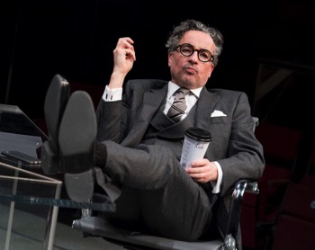 Rick (Aidan McArdle) sits reclined in an leather office chair with his feet up on the edge of the imposing glass desk, soles to the camera. He's wearing a grey wool three-piece suit with a grey striped tie. He's holding a Starbucks cup with his name on the side.