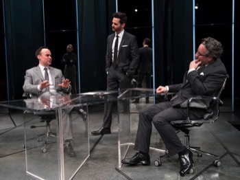 Rick (Aidan McArdle) sits a leather office chair across the glass desk from Jeff (Joseph Balderama) who's making a pitch. Seth (Tom Riley) stands at the end of the desk. Behind them are mirrored toblerone shaped panels with glowing white light in the centre of each.