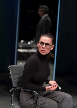  Jenny (Hayley Atwell) sits in an leather boardroom swivel chair. She wears a black turtle-neck and horn rimmed glasses. Her hair is tied back in a ponytail. Behind her is a large mirrored panel with white LED shining out at the sides. A reflection of a man in a suit is just visible. 