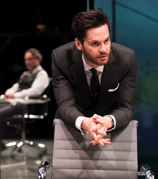 Seth (Tom Riley) leans on the back of a leather swivel chair. In the background Rick (Aidan McArdle) is just visible sat in another chair.