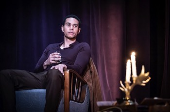  James (John Macmillan) sits in an armchair with a tumbler of whisky in his hand. His looks ahead to his left. In the foreground is a candelabra in the shape of antlers with two lit candles in. There are purple drapes behind him and it is dimly lit. 