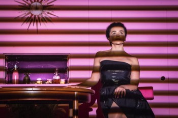  The woman (Hayley Squires) is sat at a table, facing directly towards the audience. She is wearing a black shiny shoulderless dress. Her legs are crossed and a hand is on her knee. She looks confident and knowing, with a slight smirk on her face. The room is bright pink with a recessed drinks cabinet in the wall behind, the back of which is pink mirrored, with a few decanters and seltzer bottle in front. The sole light looks like it is coming through venetian blinds in front of her so only bars of light are shining across the stage, one strip of light is illuminating just her eyes. 