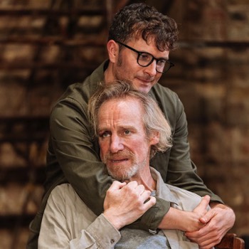  James Ekdal (Edward Hogg) hugs his father (Nicholas Farrell) from behind. His Father is sat in a chair, and holds James' arms. 