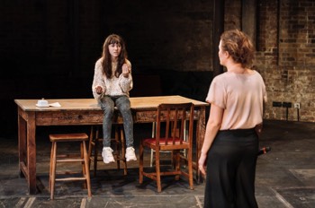 Hedwig (Clara Reed) sits on the large wooden dining table talking to her mother Gina (Lyndsey Marshal). Gina has her back to the camera. The dining table is completely clear bar a single butter dish.