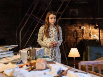  Hedwig (Grace Doherty) stands looking worried beside a table piled with books along with the left overs from their supper of bread and butter. There's also a wrapped present on the table. She's wearing paisley patterned buttoned up pyjamas. Behind her is a large wheeled painting frame ladder. A angled lamp is shining across the exposed brick wall behind. It's generally quite dark in the room. 