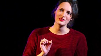  Phoebe Waller-Bridge as Fleabag. A head and shoulders image of her in a red knitted jumper. Her hair is tied back and her right hand is raised. 