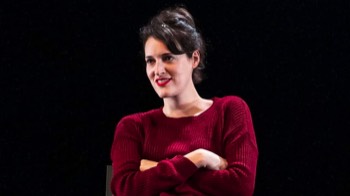 Phoebe Waller-Bridge as Fleabag. She is sat with her arms folded. Smiling looking to the side of the camera. She wears a red knitted jumper.