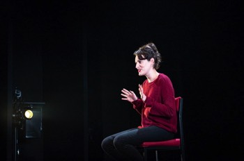  Phoebe Waller-Bridge as Fleabag. She is sat on a high stool. Her hands are up gesticulating outwards. The view is from her left side, and the camera is looking into the opposite wing, where a light pointing at her is visible. 