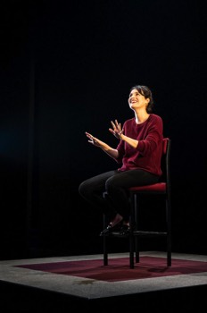  Phoebe Waller-Bridge as Fleabag. She is sat on a high stool. Her hands are up gesticulating outwards. The camera is out front of her, but lower from her so we can see the floor under her stool. She is looking upward smiling. 