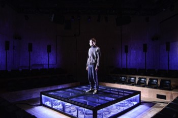  Ivan (Alex Austin) stands in the centre of the square stage. The black mesh stage platform is underlit in a cold white glowing outwards. Ivan is facing to the left, he is lit tightly in a cold white light from the left. The speakers standing behind the audience have blue light glowing out from behind them. 