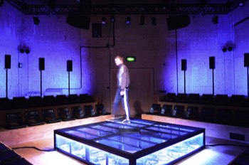  A long exposure photo of Ivan (Alex Austin) in the centre of the square stage walking to the left. The black mesh stage platform is underlit in a cold white glowing outwards, with coiled cables all over the floor under the mesh. A bright cool blue light is glowing out from behind the tall speakers behind the audience banks, so the wood panels of the walls are lit brightly. 