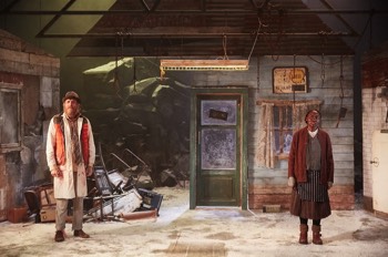  John Daniels (Rhys Ifans) and Noni (Rakie Ayola) stand wide apart in their broken down butchers shop. The floor is covered in snow, there's no roof except for some exposed beams and a portion of the back wall is missing revealing snow covered rocks behind with a stormy sky surround behind. John Daniels wears his butchers jacket under a gilet and multiple under layers. Noni wears old fashioned brown dress with a tabard over the top. She has multiple jumpers and cardigans on with a red canvas jacket over the top and knitted sleeves pulled through over her hands. 