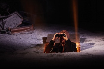 Ifan William (Sion Daniel Young) peeks out of a trap door in the floor looking through binoculars at something overhead. The trapdoor is surrounded by snow and a warm light streams out from below. Ifan wears a knitted wool hat and wears a black wool trench coat.