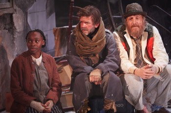 The Captain (Jason Hughes) sits between John Daniels (Rhys Ifans) and Noni (Rakie Ayola). The Captian wears a black military full length jacket with a belt at the waist, with an attached revolver holster. A very long brown fabric scarf is wrapped many times around his neck, and he has fabric wrapped around the tops of his boots. John Daniels wears his butchers jacket under a gilet and multiple under layers. Noni wears old fashioned brown dress with a tabard over the top. She has multiple jumpers and cardigans on with a red canvas jacket over the top and knitted sleeves pulled through over her hands. Behind them is a pile of broken chairs of varying types and a broken white tiled wall. All three are covered in flakes of snow.