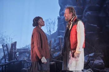John Daniels (Rhys Ifans) and Noni (Rakie Ayola) stand facing each other in front of large snow covered rocks and dead foliage. John Daniels wears his butchers jacket under a gilet and multiple under layers. Noni wears old fashioned brown dress with a tabard over the top. She has multiple jumpers and cardigans on with a red canvas jacket over the top and knitted sleeves pulled through over her hands.