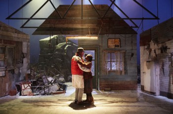  John Daniels (Rhys Ifans) and Noni (Rakie Ayola) dance together in the centre of their broken down butchers shop. The floor is covered in snow, there's no roof except for some exposed beams and a portion of the back wall is missing revealing snow covered rocks behind with a stormy sky surround behind. John Daniels wears his butchers jacket under a gilet and multiple under layers. Noni wears old fashioned brown dress with a tabard over the top. She has multiple jumpers and cardigans on with a red canvas jacket over the top and knitted sleeves pulled through over her hands. 
