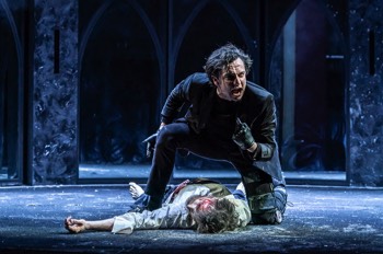  Richard III (Tom Mothersdale) kneels over the body of Henry, who is bleeding from his head. Richard is wearing all black with black leather gloves. He has an articulated brace on his left leg. He's pointing at himself and angrily shouting. A curved wall of arched mirrors sits at the back of the stage, the floor looks like black gravel. 