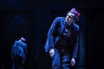 Richard III (Tom Mothersdale) stands hunched wearing a pink paper party hat. His left leg is in an articulated brace. The reflection of his back is visible in a mirrored arched panel behind him.