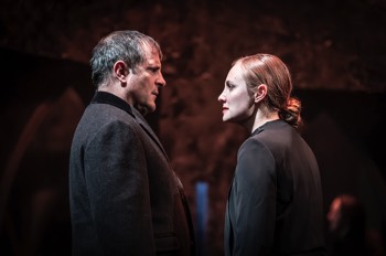  Norfolk (Leila Mimmack) confronts Stanley (Michael Matus). They stand close facing each other, with their sides to the camera. Both are wearing black jackets. The light is warm and orange. 