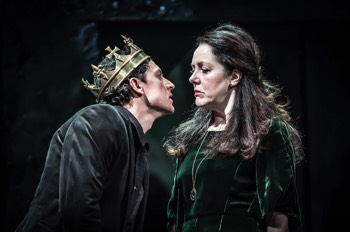 Richard III (Tom Mothersdale) gets up in the face of Elizabeth (Derbhle Crotty). Elizabeth looks down her nose at him. She wears a green velvet dress and a long gold thin necklace with a gold droplet on the end. Richard is wearing a dull gold metal crown.