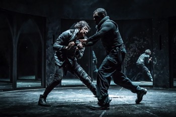  Richard III (Tom Mothersdale) battles Richmond (Caleb Roberts). Richmond has grabbed Richard's forearm as Richard tries to stab Richmond with a knife. Richmond wears black combat trousers, and a black stab vest. Richard is in a black jacket with his left leg in an articulated leg brace. 