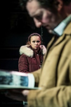  Tim (John Hollingworth) is stood close to the camera out of focus, reading a magazine. Beyond him, we see Dani (Jessica Rhodes) sharply. She is wearing a red puffer jacket with a white fluffy edged hood, a checked school skirt is just visible below. She stares at Tim. Tim as a tan cord jacket on. Between them is a chain from a playground swing. The light is bright but cold. 