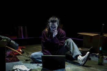  Dani (Jessica Rhodes) sits on the floor with a laptop between her legs. She's surrounded by boxes, and some left over bowls and an empty bottle of wine. The 90s style Dell laptop is plugged in to the edge of the stage, which is illuminated in a dim purple LED surround. It's dark and she's lit very dimly. She is chewing a fingernail and look aprehensive. 