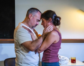 A Fight Against Production Image. Carla (Jimena Larraguivel) and Alejandro (Joseph Balderama) hold each other in front of their kitchen table. There the light of a anglepoise lamp on the wall behind them.